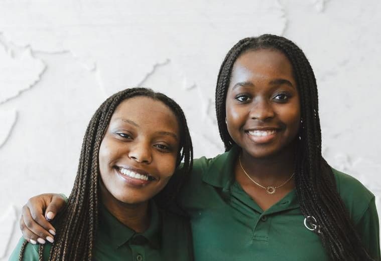 Two Greenwood Project Scholars in green Greenwood polos smiling arm in arm in front of a white relief map of the world.