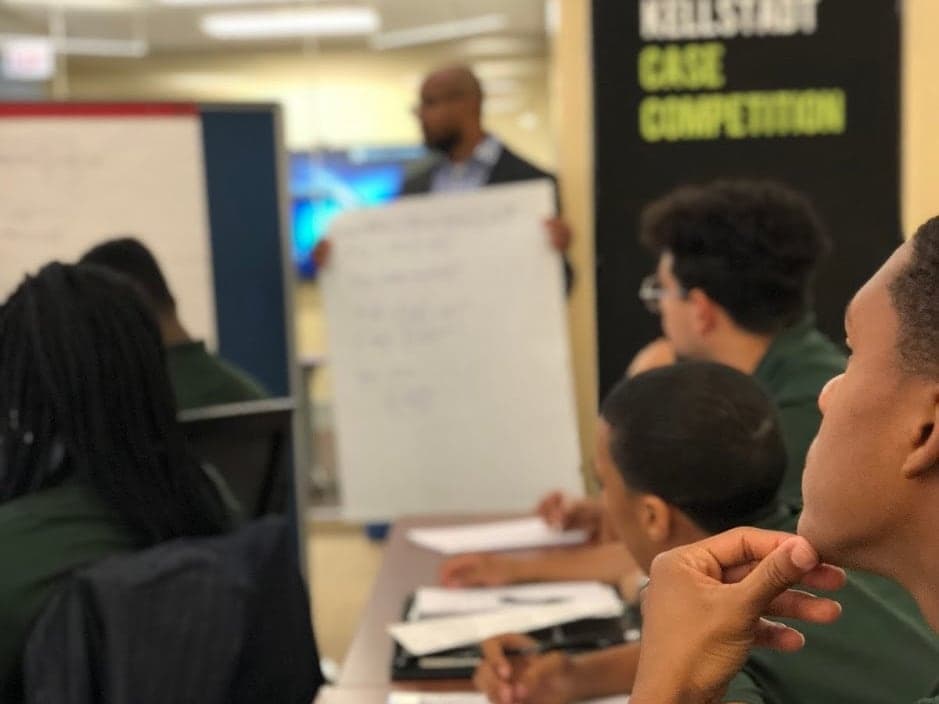 A finance professional walks Greenwood Scholars through a case at a whiteboard while they take notes.
